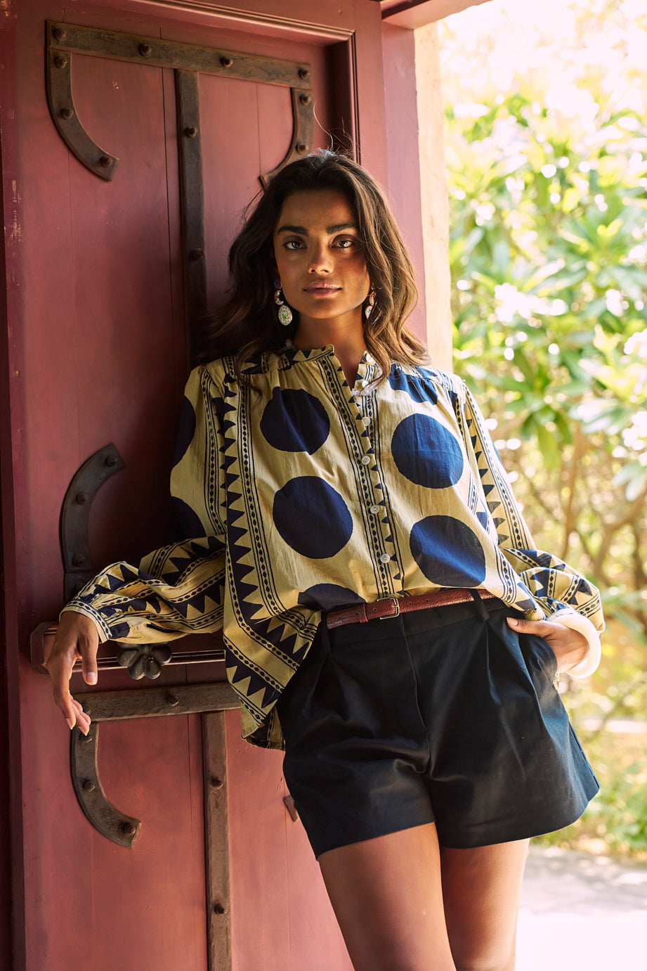 Model wearing the KLEED Midnight Geo Balloon Shirt in 100% cotton, styled with black tailored shorts and a brown leather belt, standing against a rustic red wooden door and wearing statement earrings.