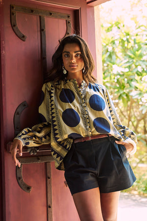 Model wearing the KLEED Midnight Geo Balloon Shirt in 100% cotton, styled with black tailored shorts and a brown leather belt, standing against a rustic red wooden door and wearing statement earrings.