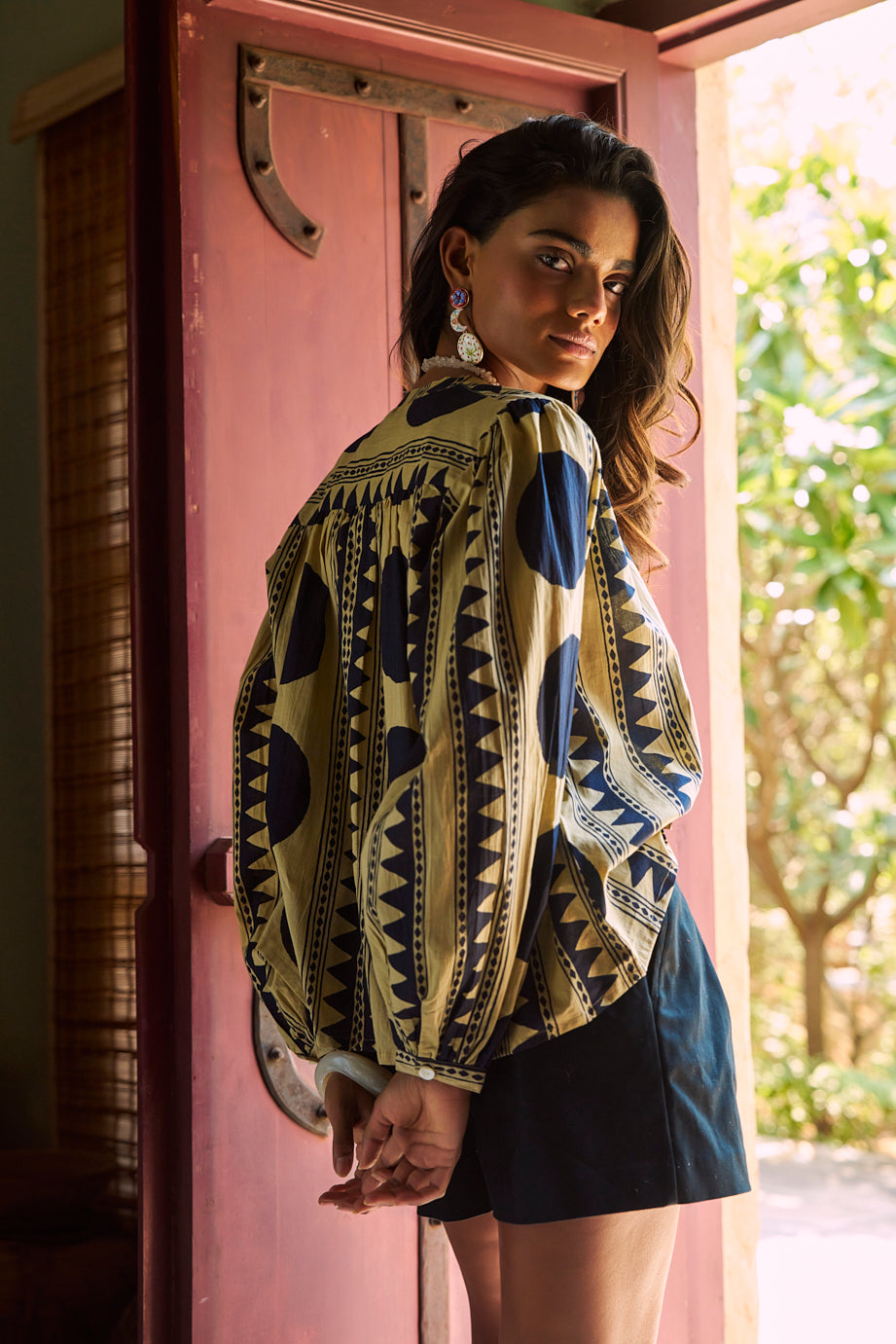 Back view of a model wearing the KLEED Midnight Geo Balloon Shirt in 100% cotton, highlighting the bold black geometric print, voluminous sleeves and relaxed fit, styled with black tailored shorts and colourful statement earrings by a rustic red wooden door.