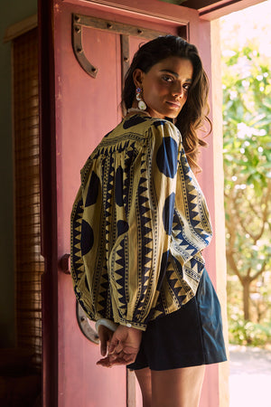 Back view of a model wearing the KLEED Midnight Geo Balloon Shirt in 100% cotton, highlighting the bold black geometric print, voluminous sleeves and relaxed fit, styled with black tailored shorts and colourful statement earrings by a rustic red wooden door.