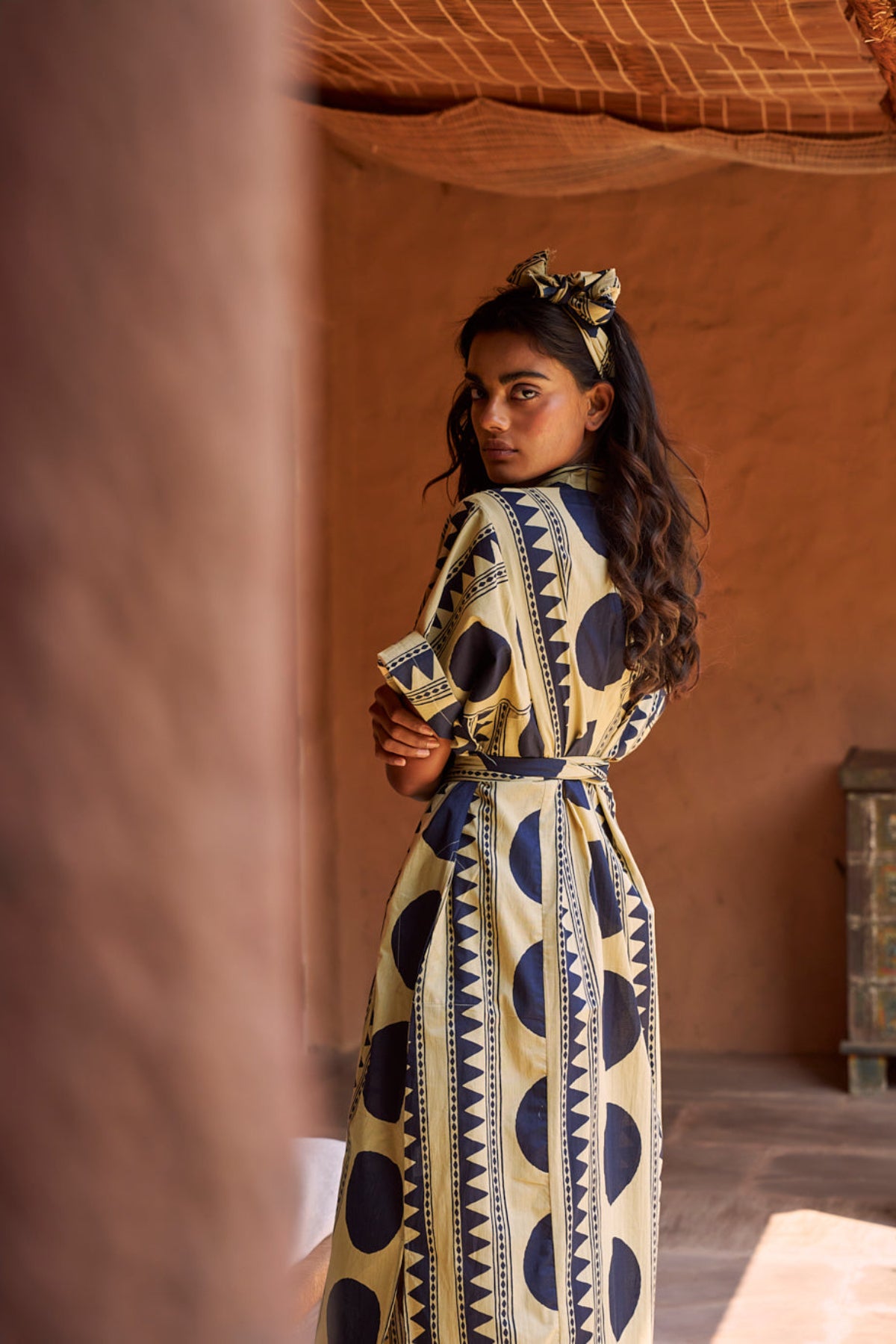 model wearing a midnight geo kaftan with terracota wall behind. Kaftan with dark blue geometric pattern on beije base and a scarf same fabric in the head. By Kleed.