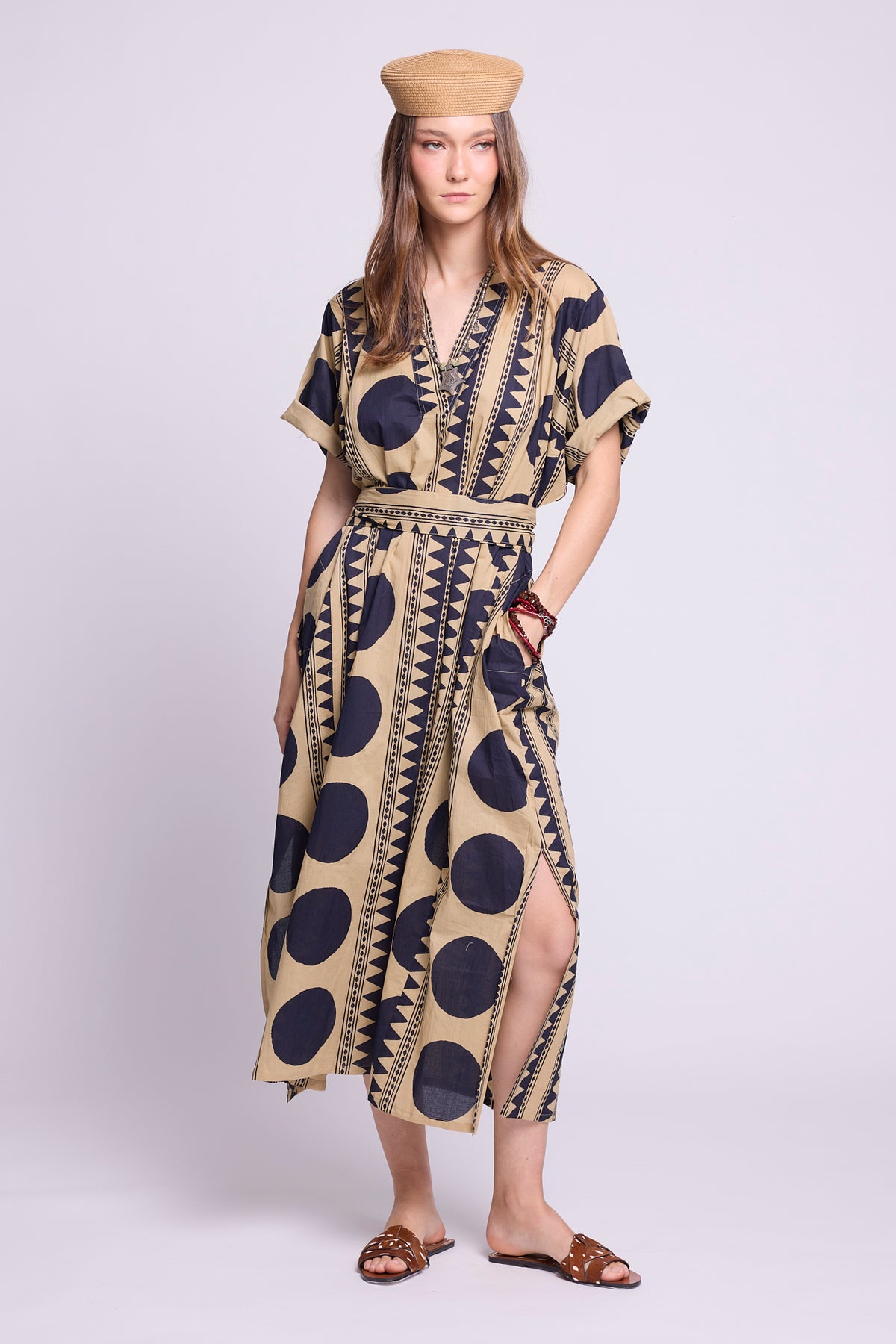 A woman modeling a beige kaftan dress with a bold navy blue polka dot and geometric pattern, paired with a matching beige beret and brown sandals, against a light gray studio background. By kleed.