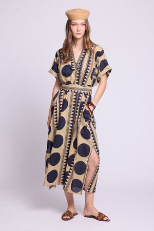 A woman modeling a beige kaftan dress with a bold navy blue polka dot and geometric pattern, paired with a matching beige beret and brown sandals, against a light gray studio background. By kleed.