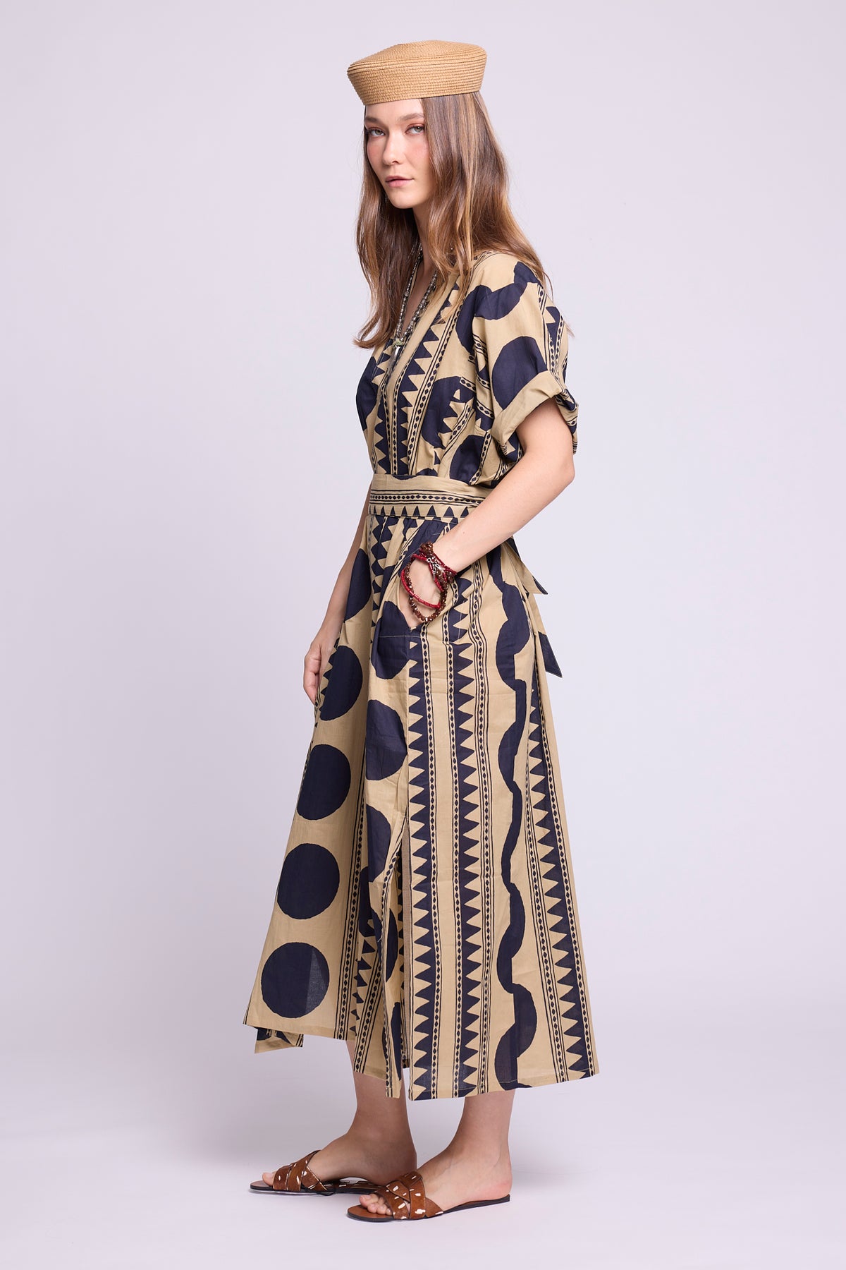 A woman modeling a beige and navy patterned midi dress with a tie waist, paired with a matching beige hat and flat sandals, against a light gray studio background. By Kleed
