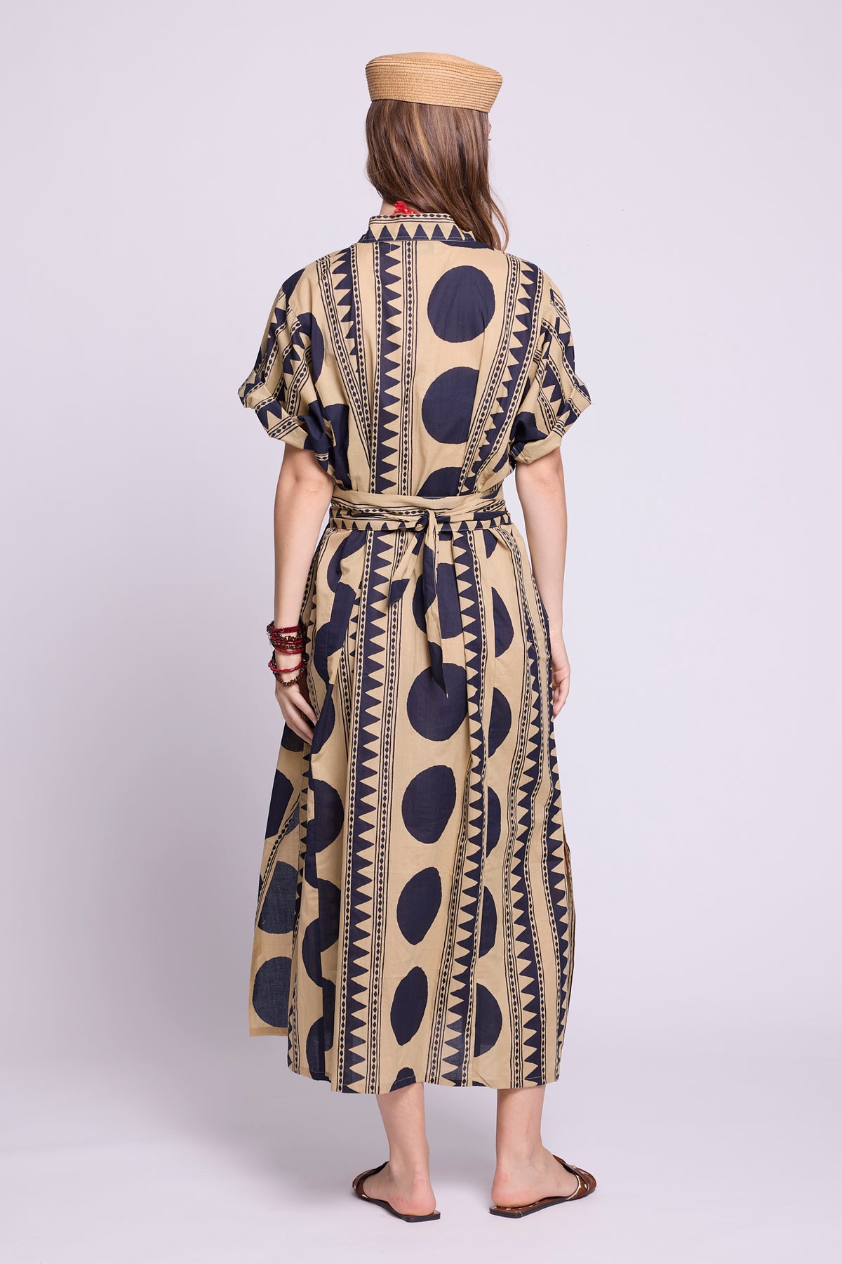 A woman viewed from the back wearing a beige and navy patterned midi dress with short sleeves, a tie waist, and a tan beret, standing against a light gray studio background. by kleed