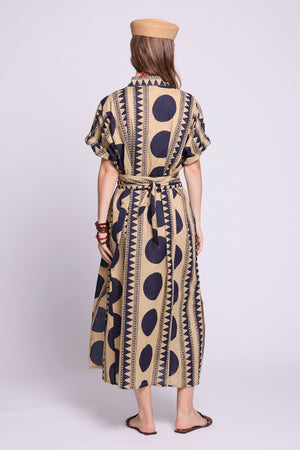 A woman viewed from the back wearing a beige and navy patterned midi dress with short sleeves, a tie waist, and a tan beret, standing against a light gray studio background. by kleed