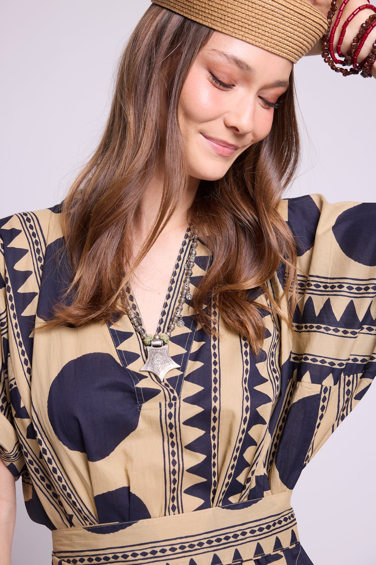 A woman wearing a beige and navy blue patterned kaftan dress with a matching belt, a woven straw hat, and red bangles, photographed against a light gray studio background. By Kleed