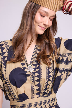 A woman wearing a beige and navy blue patterned kaftan dress with a matching belt, a woven straw hat, and red bangles, photographed against a light gray studio background. By Kleed
