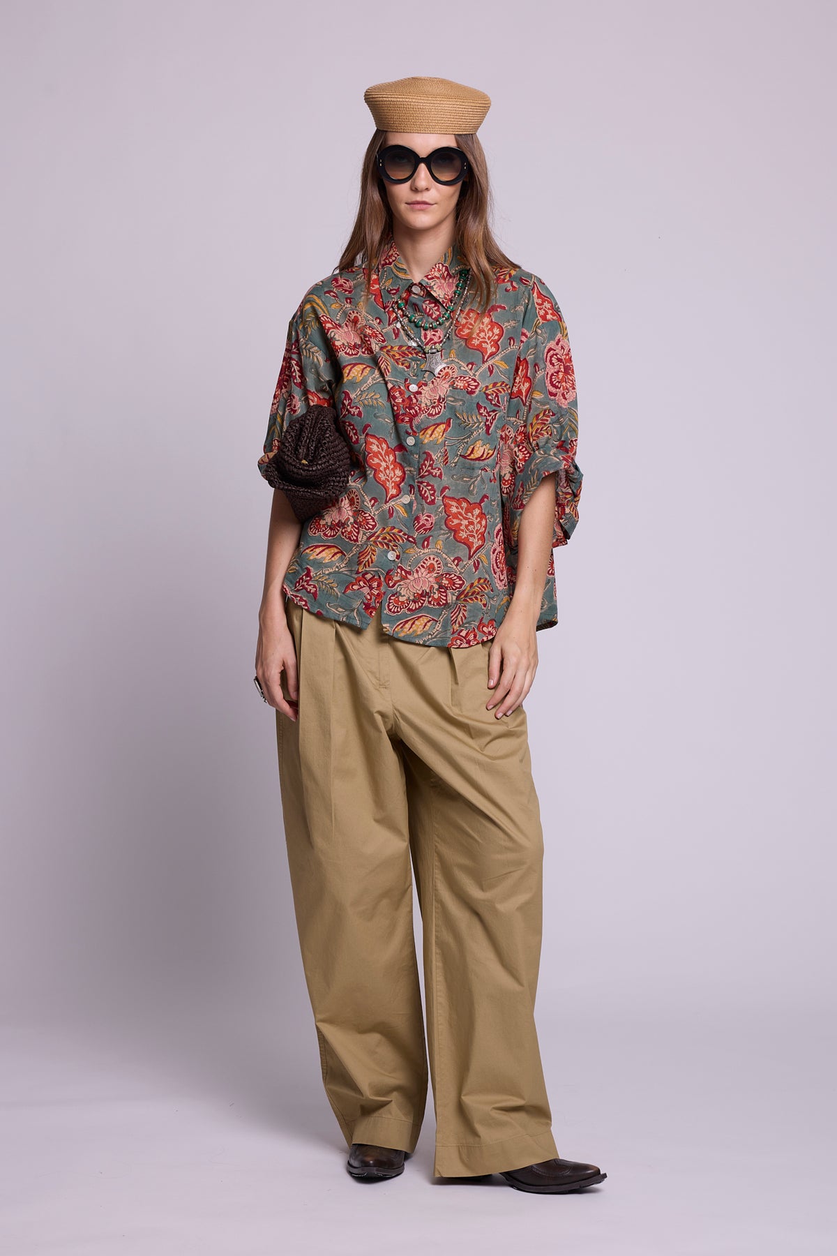Studio photo of a model wearing the KLEED Mogra Green Pyjama Shirt in 100% hand block printed cotton, styled with wide-leg khaki trousers, oversized sunglasses, a woven hat, layered necklaces and a textured dark bag.