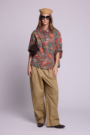 Studio photo of a model wearing the KLEED Mogra Green Pyjama Shirt in 100% hand block printed cotton, styled with wide-leg khaki trousers, oversized sunglasses, a woven hat, layered necklaces and a textured dark bag.