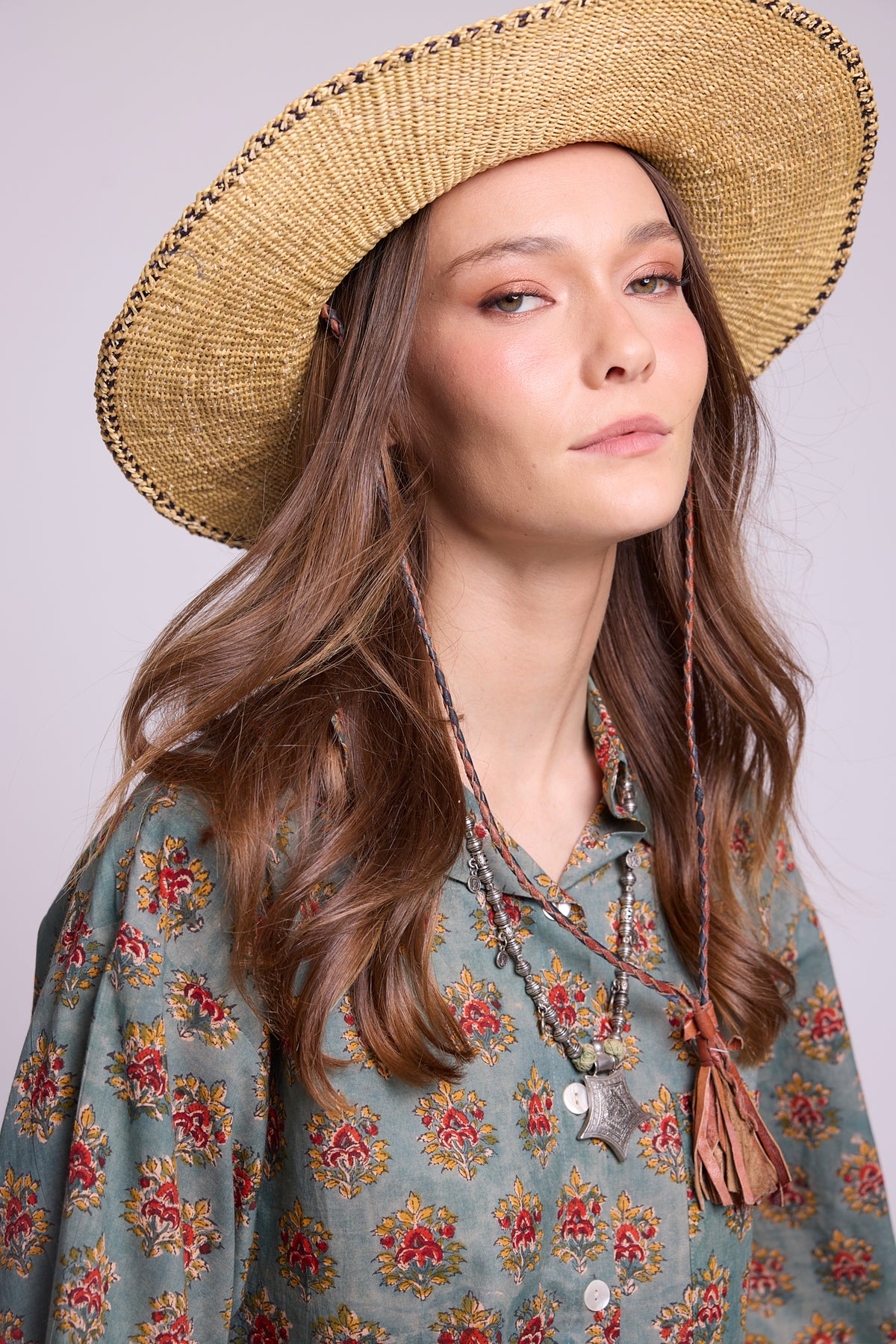 Close-up studio photo of a model in a woven straw hat wearing the top of the Mogra Hara Pyjama by Kleed, showing the red and ochre floral motifs hand block-printed on sage green cotton, collar and buttons, styled with a silver pendant and leather tassel necklace.