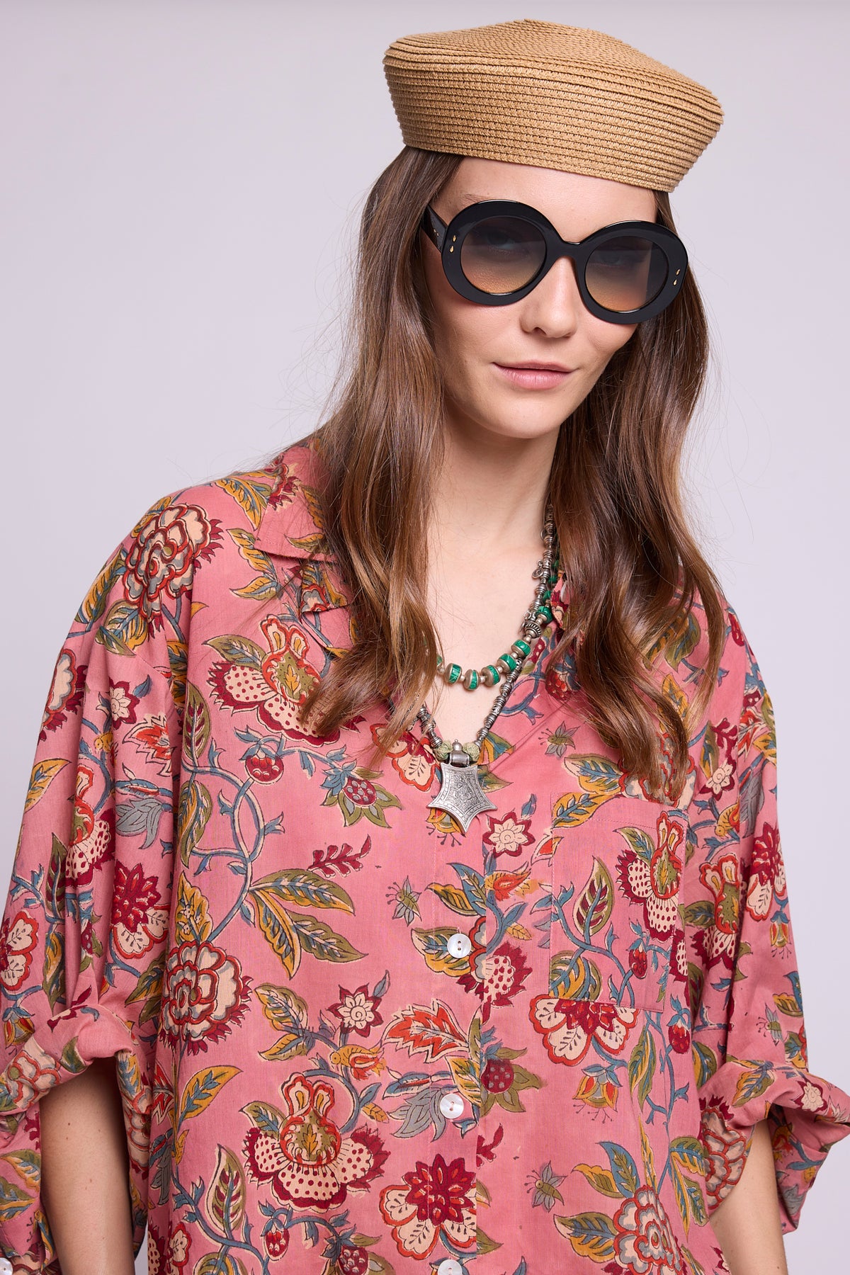 Detail studio photo of a model wearing the top of the Mogra Pink Pyjama by Kleed, showing the collar, mother-of-pearl buttons and hand block-printed floral pattern in red, green and blue on pink cotton, styled with a straw hat, black round sunglasses and a turquoise necklace with silver pendant.