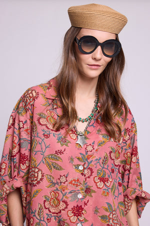Detail studio photo of a model wearing the top of the Mogra Pink Pyjama by Kleed, showing the collar, mother-of-pearl buttons and hand block-printed floral pattern in red, green and blue on pink cotton, styled with a straw hat, black round sunglasses and a turquoise necklace with silver pendant.