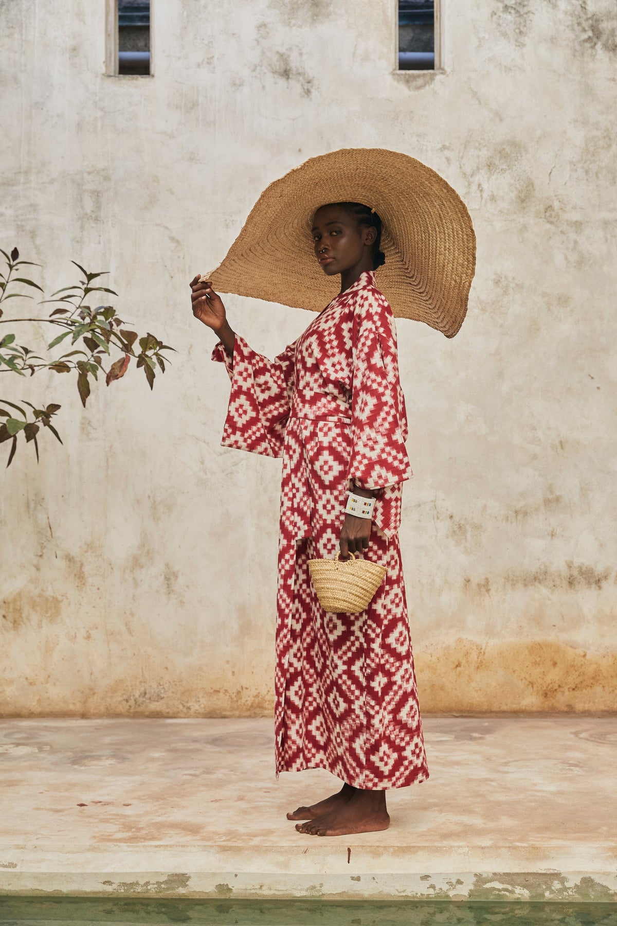 Woman in a red and kleed white geometric patterned Ikat kimono holding a straw hat and basket against a textured wall.