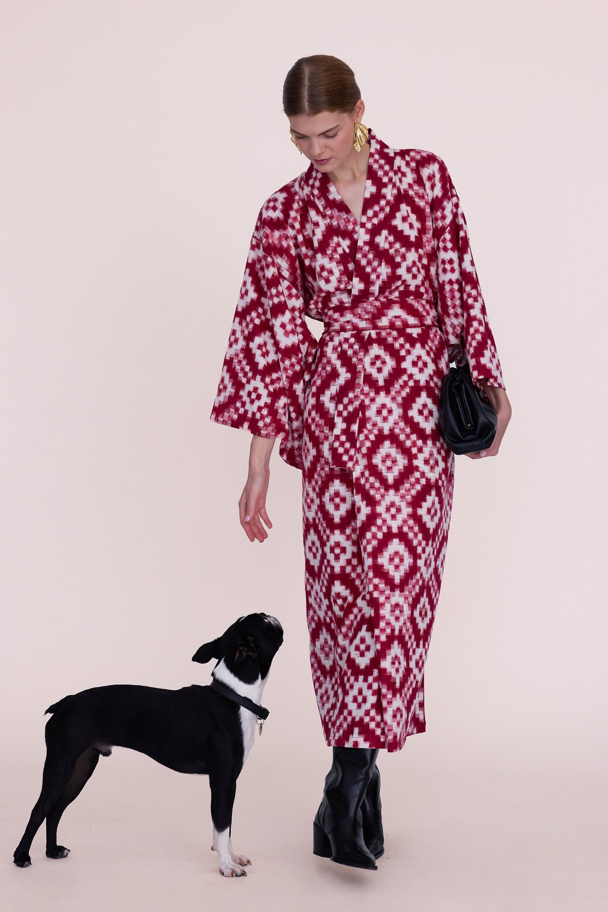 Woman wearing Mombasa red Ikat cotton kimono by Kleed with black bag and boots, standing next to a Boston Terrier dog, conscious luxury loungewear