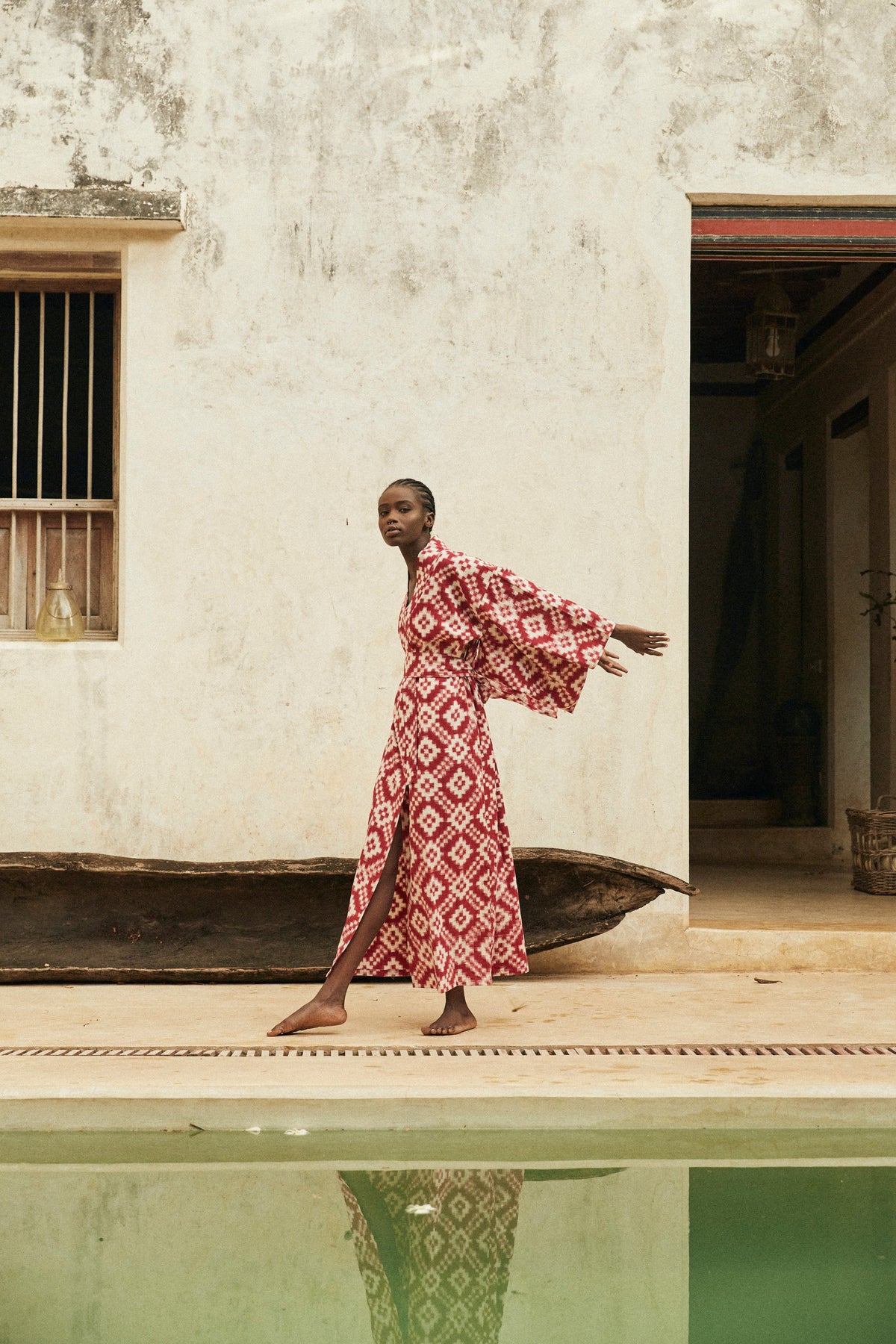 Woman wearing Mombasa red Ikat cotton kimono by Kleed in outdoor editorial campaign, standing barefoot beside a wooden boat next to a pool, sustainable luxury loungewear