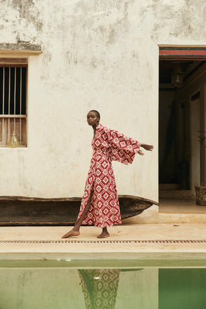 Woman wearing Mombasa red Ikat cotton kimono by Kleed in outdoor editorial campaign, standing barefoot beside a wooden boat next to a pool, sustainable luxury loungewear