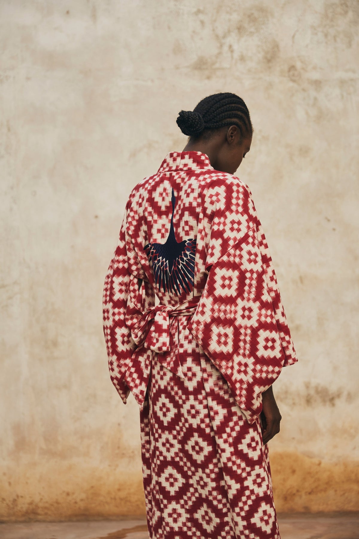 Back side of the Mombasa red Ikat cotton kimono by Kleed, hand embroidered bird detail worn in outdoor editorial campaign against rustic wall, artisan sustainable loungewear