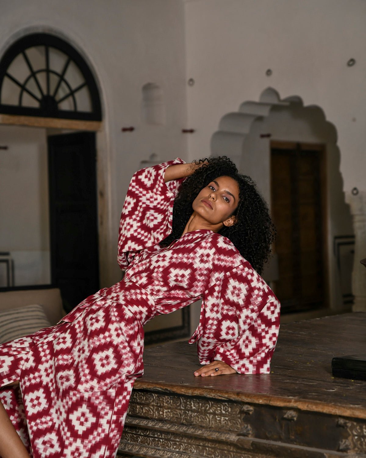 Woman wearing Mombasa red Ikat cotton kimono by Kleed posing on antique wooden table inside Indian haveli, editorial campaign shot, conscious luxury loungewear