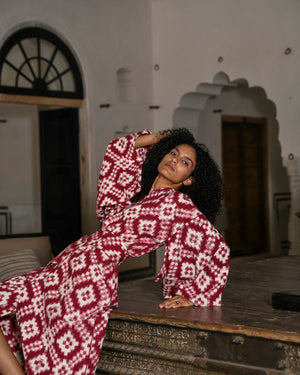 Woman wearing Mombasa red Ikat cotton kimono by Kleed posing on antique wooden table inside Indian haveli, editorial campaign shot, conscious luxury loungewear