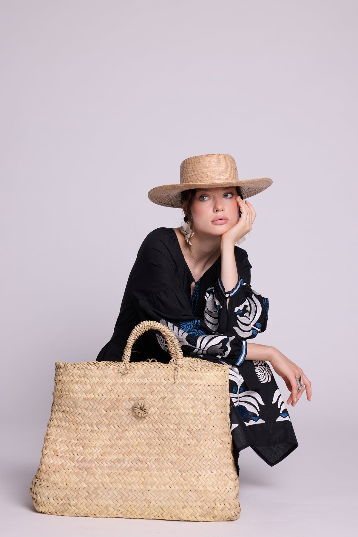 Model seated wearing the KLEED Portofino Black Kaftan with white embroidered details, blue tassels and a woven straw bag.