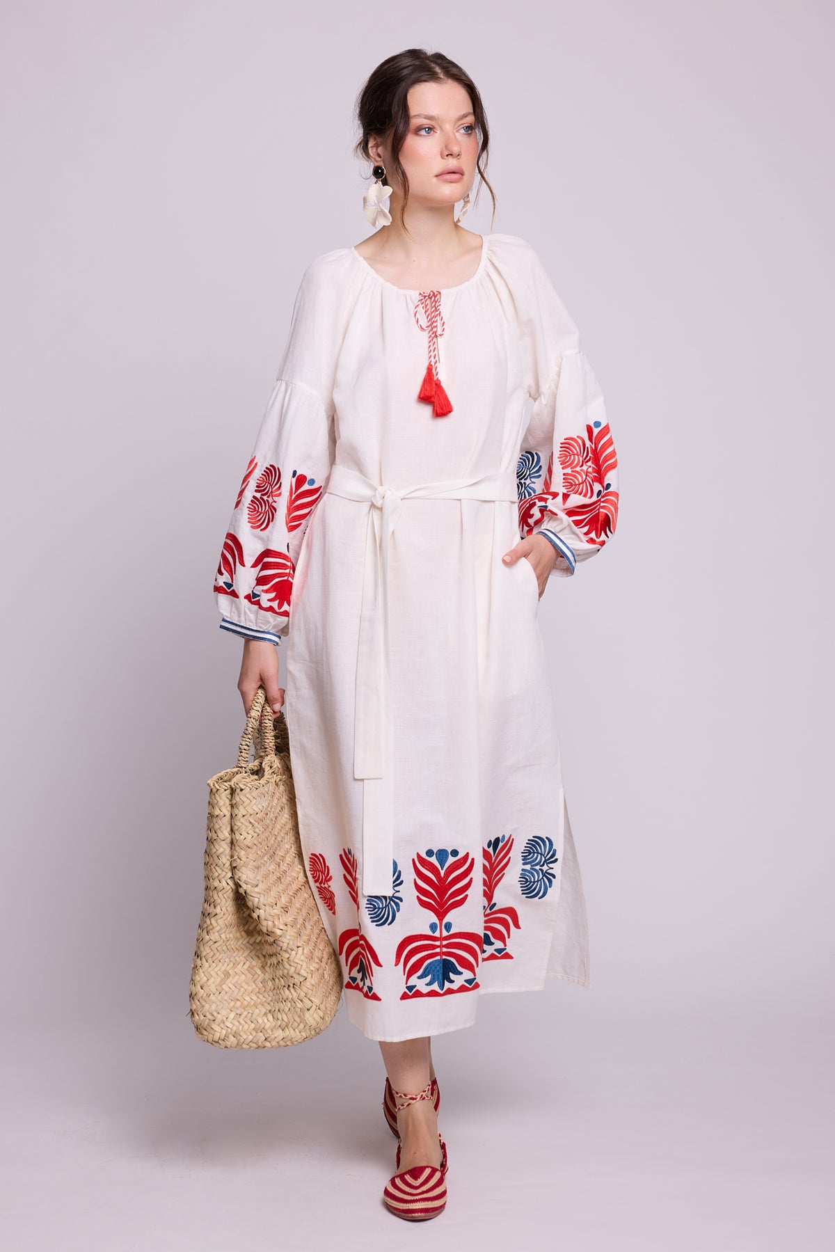 Model seated wearing the KLEED Portofino Black Kaftan with white embroidered details, blue tassels and a woven straw bag.