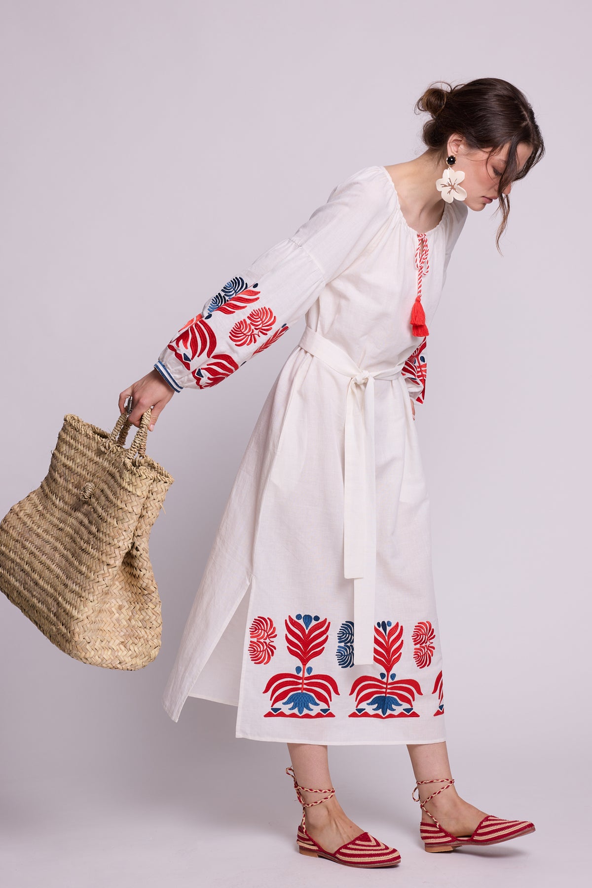 Side view of model wearing the KLEED Portofino White cotton-linen Kaftan with red and blue embroidered sleeves and hem, statement earrings, red sandals and a woven straw basket.