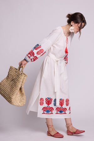 Side view of model wearing the KLEED Portofino White cotton-linen Kaftan with red and blue embroidered sleeves and hem, statement earrings, red sandals and a woven straw basket.