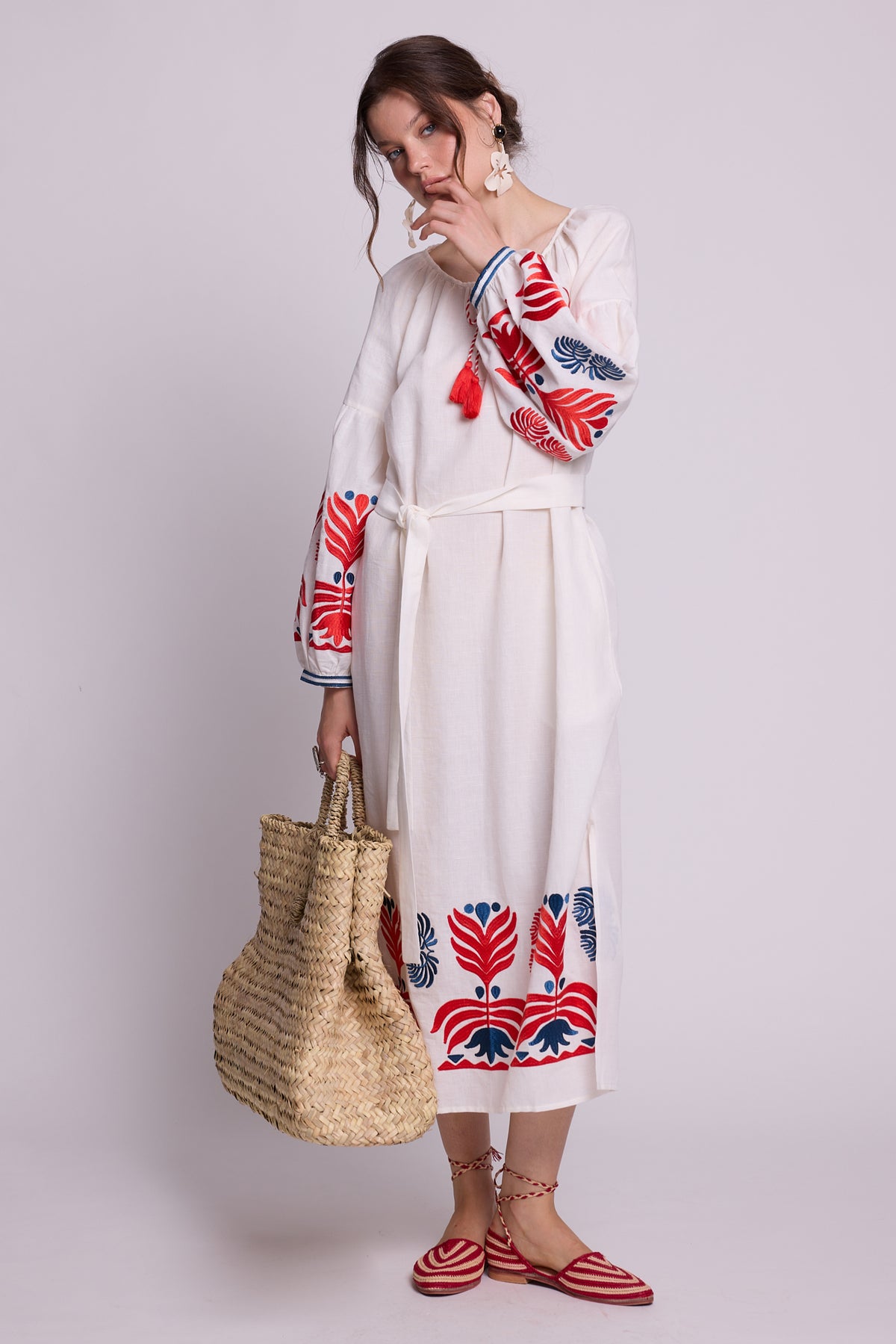 Full-length model wearing the KLEED Portofino White Kaftan in cotton linen, featuring red and blue embroidery, styled with earrings, red flat sandals and a large woven straw basket.