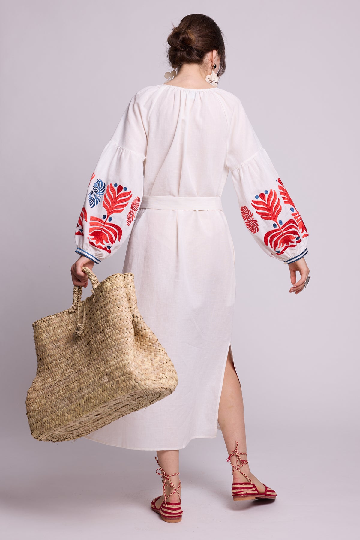 Full-length model wearing the KLEED Portofino White Kaftan in cotton linen, featuring red and blue embroidery, styled with earrings, red flat sandals and a large woven straw basket.