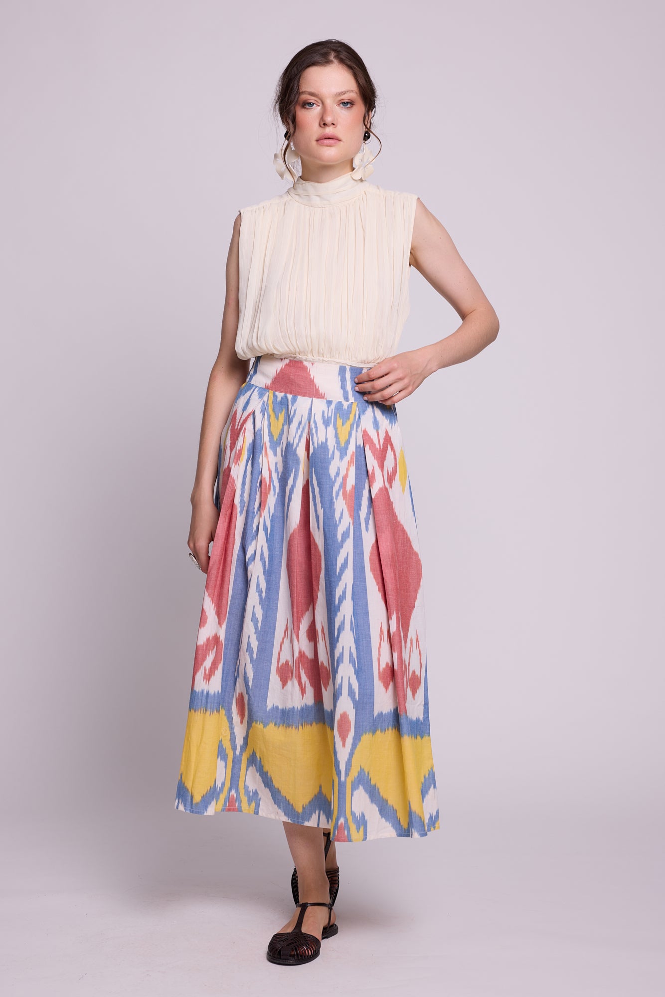 A model wearing a sleeveless cream pleated top and the seashore skirt by kleed, a high-waisted midi skirt with a colorful blue, pink, yellow, and white ikat pattern, standing against a light gray studio background.