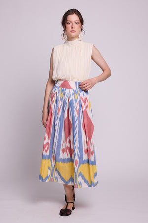 A model wearing a sleeveless cream pleated top and the seashore skirt by kleed, a high-waisted midi skirt with a colorful blue, pink, yellow, and white ikat pattern, standing against a light gray studio background.