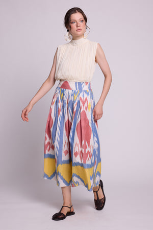 A model wearing a sleeveless cream pleated mock-neck top and the seashore skirt by kleed, a high-waisted midi skirt with a colorful blue, pink, and yellow ikat pattern, paired with black flat shoes, against a light gray studio background.