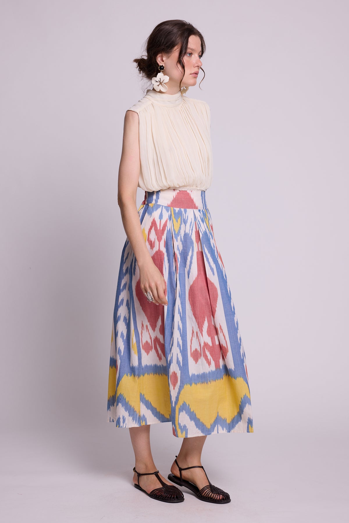 side view of a woman modeling a sleeveless cream pleated top and the seashore skirt by kleed, a high-waisted midi skirt with a colorful ikat pattern in blue, pink, and yellow, paired with black strappy flat sandals against a light gray studio background.