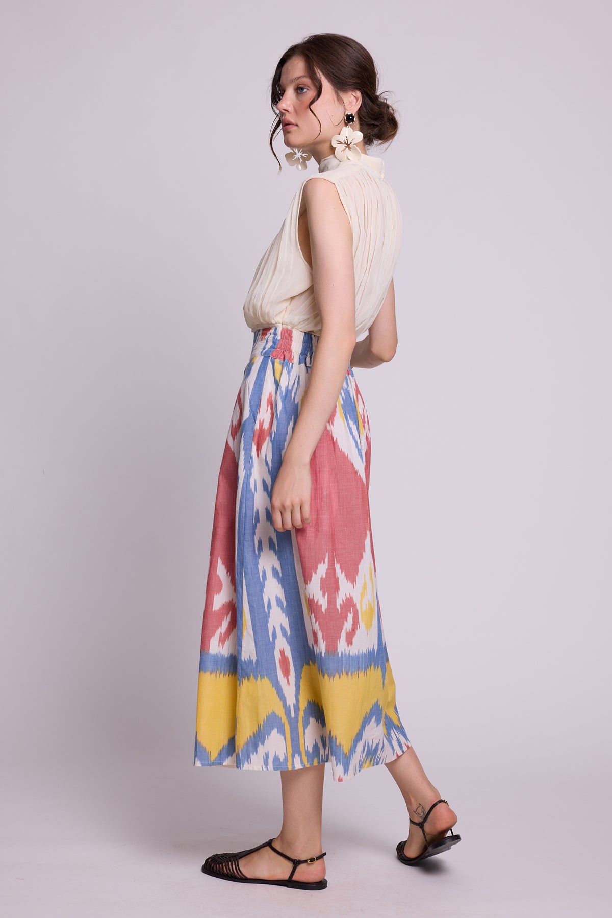 A woman modeling a sleeveless cream top and the seashore skirt by kleed, a high-waisted midi skirt with a colorful ikat pattern in pink, blue, yellow, and white, paired with black strappy sandals against a light gray studio background.