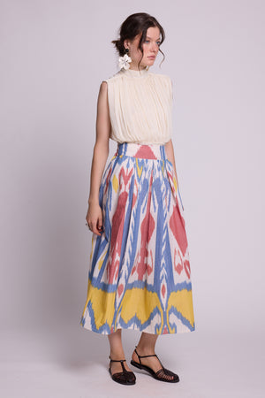 A woman modeling a sleeveless cream pleated top and a the seashore skirt by kleed a high-waisted midi skirt with a colorful ikat pattern in blue, red, yellow, and white, paired with brown strappy sandals against a light gray studio background.